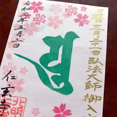 
私たち真言宗では、インド由来の聖なる文字・梵字をとても大切にしています。

どのくらい大切にしているかといいますと、「単なる記号でも名前でもなく、『仏さまそのもの（現身）』である」と考えるほどです。
　
初めての「とてもカラフルなご朱印」ということで、「ご朱印とは何か？」「ご朱印で最も大切なものは何か？」を真剣に考えましたが、【仁玄寺としては、梵字だけは外せない】という結論に落ち着きました。
　
それからも、さまざまな試行錯誤しましたが、最終的に弘法大師・空海さんの梵字「ユ」字を中央に置き、他のハンコや文字を被せないことにしました。
　
空海さんの現身として、皆さまをお守りくださることと思います。どうか、大切にお持ちくださいね。

◎金文字の「御入定（ごにゅうじょう）」と桜への思い
